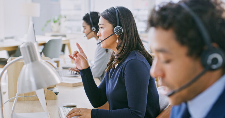 Call center agent wearing a headset and sitting in front of a computer. Other contact center agents can be seen in the background.