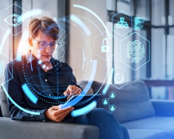 Serious young European businessman using tablet computer sitting on sofa in blurry office with double exposure of immersive facial recognition interface. Concept of biometric scanning